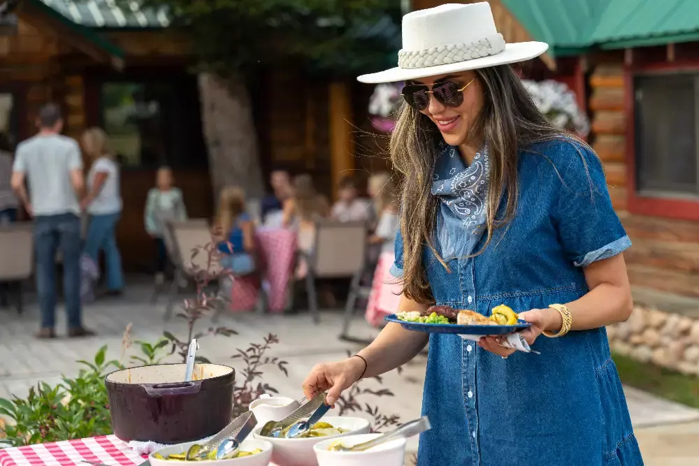 A woman in a hat and sunglasses serving herself food from a table. The table is filled with dishes and utensils