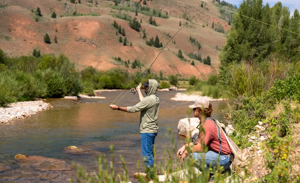 Two people by a river, one holding a fishing rod and the other holding a camera