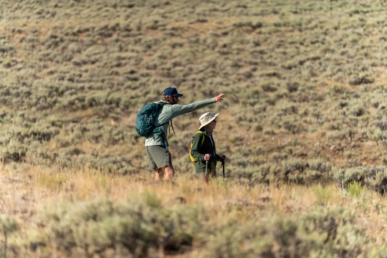 Two people in a field with backpacks and one pointing to something in the distance