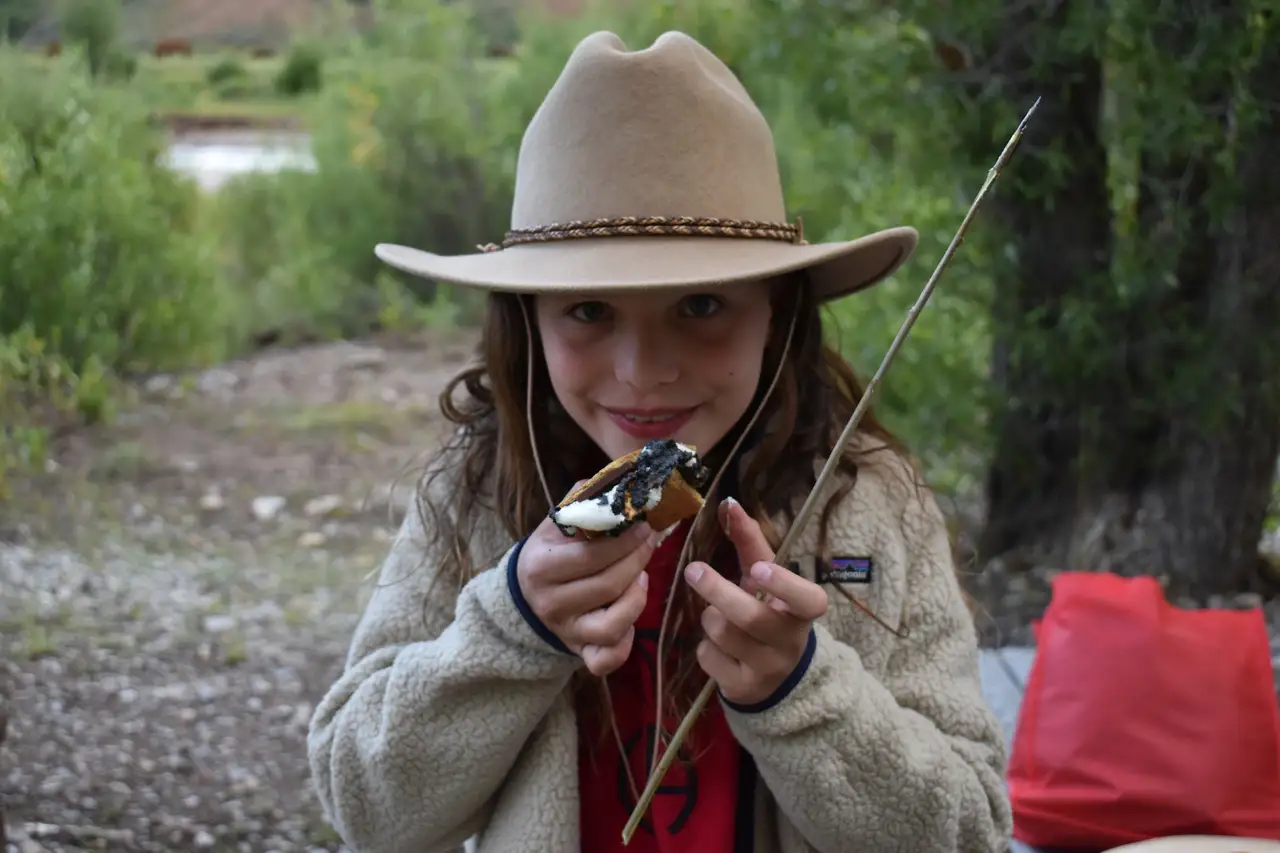 A girl eating a snack while holding a fishing rod and wearing a cowboy hat