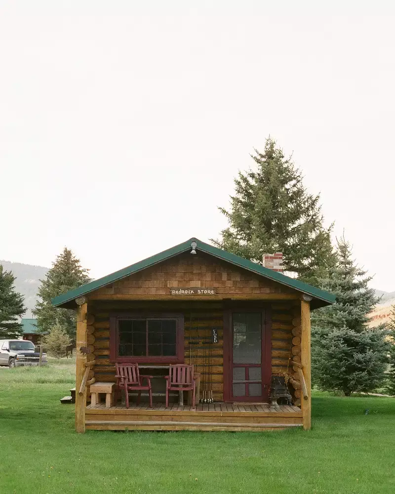 A small log cabin with a green roof and red trim. The cabin has a deck with a bench and a picnic table. The cabin is surrounded by trees and has a car parked nearby