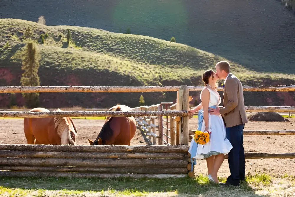 A couple kissing in front of a fence with a horse in the background
