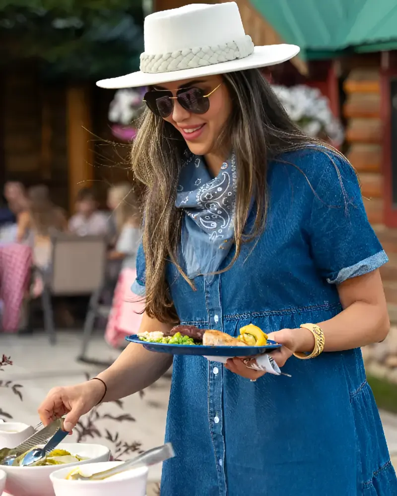 A woman wearing a hat serves food at a picnic table, surrounded by a cheerful outdoor setting